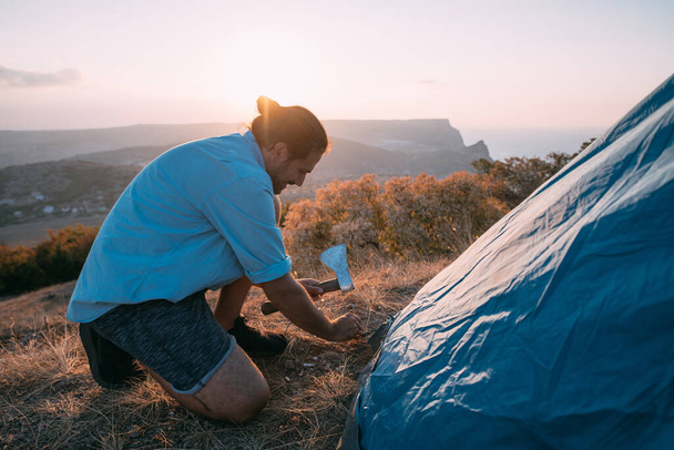 Young male tourist puts a tent in the mountains. The guy sets up a camp on a hike, collects a tent alone in the mountains at sunset - Photo, Image