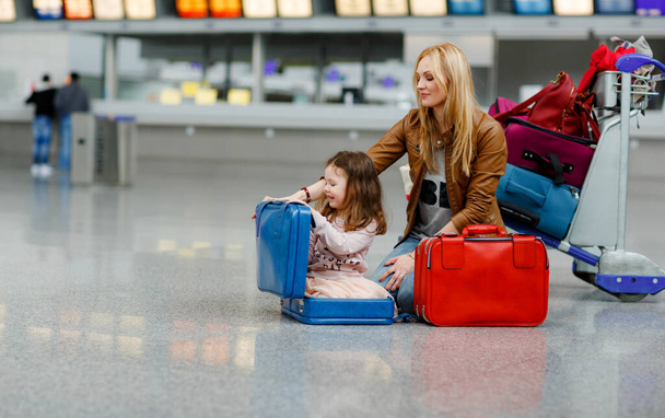 Cute little girl and mother at the airport. Tired family sitting on ground and waiting for delayed flight. Woman and child by luggage trolley with suitcases at terminal. Travel lifestyle - Foto, afbeelding