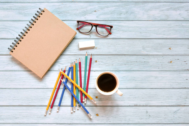 Top view of brown closed cover blank notebook with coffee cup with eyeglasses and various colorful pencil on wood table background. Flat lay, creative workspace, Business office or education concept - Photo, Image