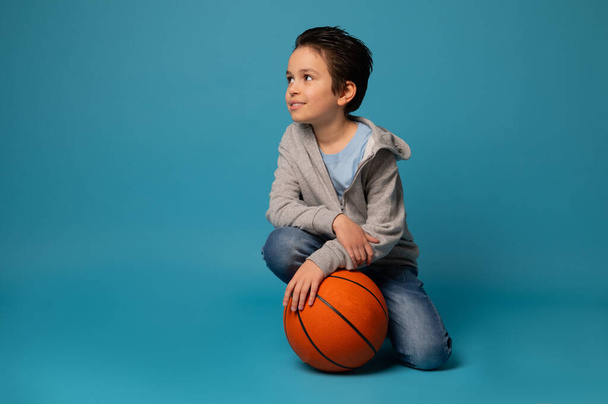 Beautiful sporty teenage boy posing with a ball for playing basketball over blue background. Concept of healthy and active lifestyle on blue background with copy space - Photo, Image