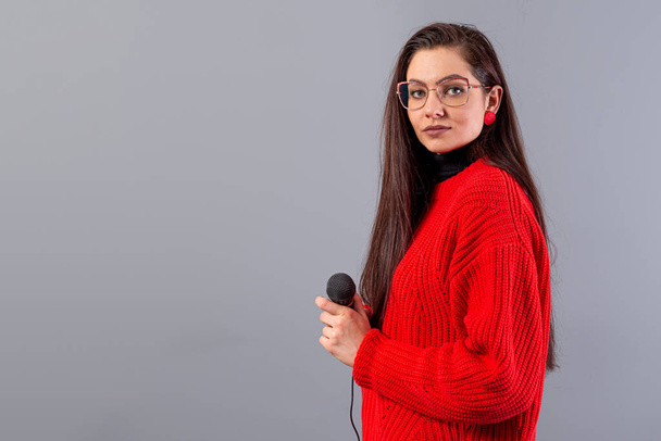 young, emotional brunette with a microphone dressed in a red sweater sings karaoke or says a speech, isolated on gray - Fotografie, Obrázek