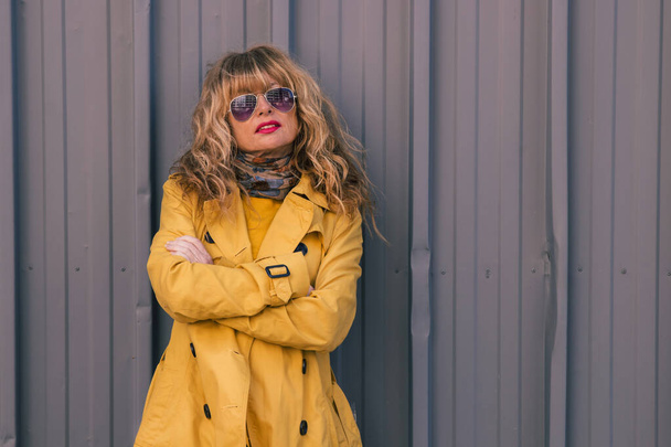 atrakcyjna blondynka w okularach przeciwsłonecznych i żółtym płaszczu na ścianie ulicy - Zdjęcie, obraz