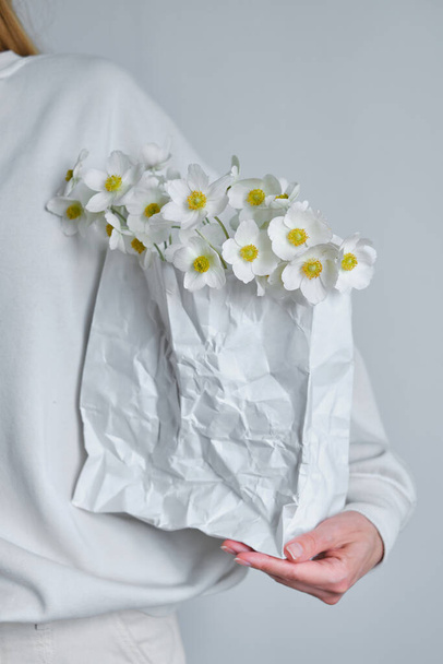 Anemones in a white craft bag in the hands of a happy woman. The florist girl gathered a bouquet. Beautiful spring white flowers. Gift for the holiday, celebration of the spring mood. Fresh floral - Fotografie, Obrázek