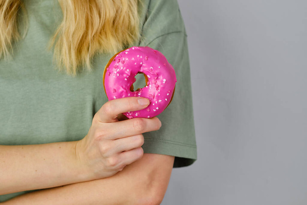 Pink doughnut in the hands of a girl. Delicious sweet american dessert. Popular baked goods. Donut in bright sugar glaze close-up. Woman posing with donut in her hands. Fast food - Photo, image