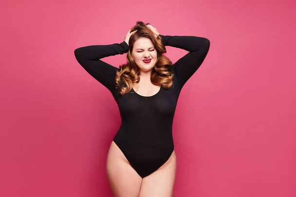 Beautiful emotional plus-size female model in a black bodysuit posing on pink background in studio - Fotografie, Obrázek