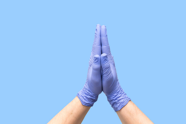 Die Hände in medizinischen Latexhandschuhen, die zusammengefaltet sind, weisen auf den Prozess des Gebets hin. Lasst uns über eine schnelle und einfache Behandlung beten. Gute Reinigung. Palmen isoliert auf blauem Hintergrund. Nahaufnahme. Banner. Hoffnung in der Medizin. - Foto, Bild