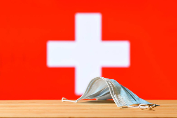Μια ιατρική μάσκα βρίσκεται στο τραπέζι με φόντο τη σημαία της Ελβετίας. Η έννοια του υποχρεωτικού καθεστώτος μασκών για τους κατοίκους της χώρας και τους τουρίστες στην Ελβετία κατά τη διάρκεια πανδημίας. - Φωτογραφία, εικόνα