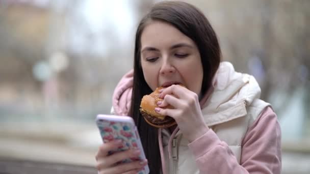 Счастливая женщина ест бургер и использует смартфон на улице - Кадры, видео