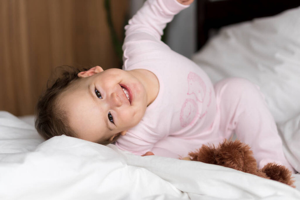 Authentic portrait caucasian little infant chubby baby girl or boy in pink sleepy upon waking looking at camera smiling cute and grimaces in white bed. Child care, Childhood, Parenthood, life concept - Photo, image