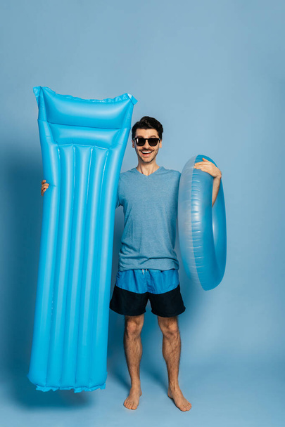 Full-length of amazed happy caucasian shirtless guy, standing with blue inflatable ring and mattress on an isolated blue background in sunglasses looks at camera and smiling, vacation concept - Fotó, kép