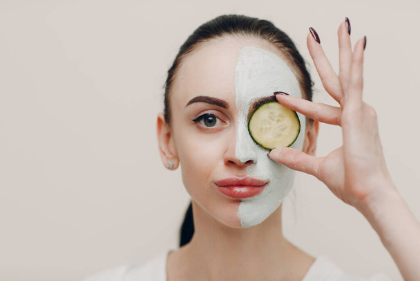 Jovem mulher aplicando máscara de barro no rosto com pepino em olhos em spa de beleza - Foto, Imagem