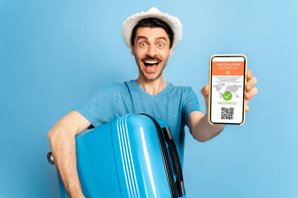 Electronic certificate of vaccination of coronavirus infection covid19.Defocused guy holds smartphone with electronic certificate of vaccination and suitcase in hands,stand on isolated blue background - Photo, Image
