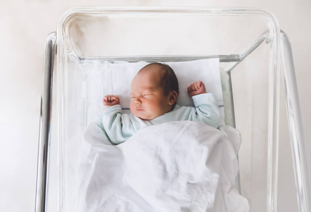 Newborn baby is sleeping in small transparent portable plastic bed. Baby first days of life is lying in a hospital crib after birth. - Фото, зображення