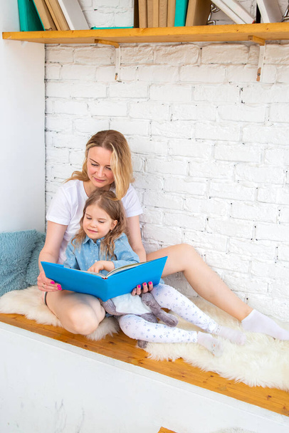 Happy caucasian mother with her daughter reading a book and smiling while sitting on the floor in the living room - Фото, зображення