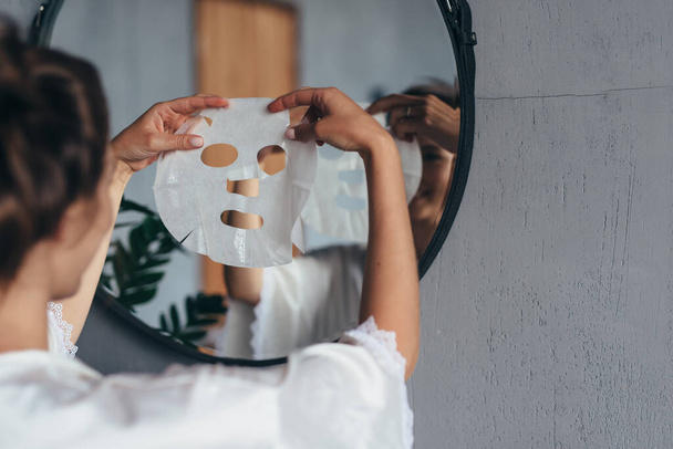 Woman with sheet mask in her hands in the bathroom before applying it to her face - Foto, afbeelding