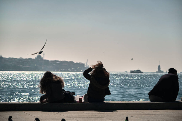 Τουρκία istanbul 04.03.2021. Κορίτσια που κάθονται στην όχθη του αφεντικού. σγουρά μαλλιά γυναίκες και τις τρίχες της κύματα από ανέμους με γλάρους, πλοίο και πλοίο φόντο κατά τη διάρκεια του πρωινού και τον ήλιο αντανάκλαση στη θάλασσα. - Φωτογραφία, εικόνα