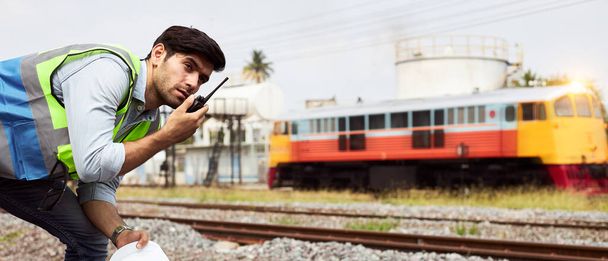 Pomyślni specjaliści ds. logistyki kolejowej lub inżynierowie kolejowi trzymający kaski i noszący kamizelki ochronne stojące na zewnątrz toru kolejowego posiadają krótkofalówkę sprawdzającą gotowość do pracy. - Zdjęcie, obraz