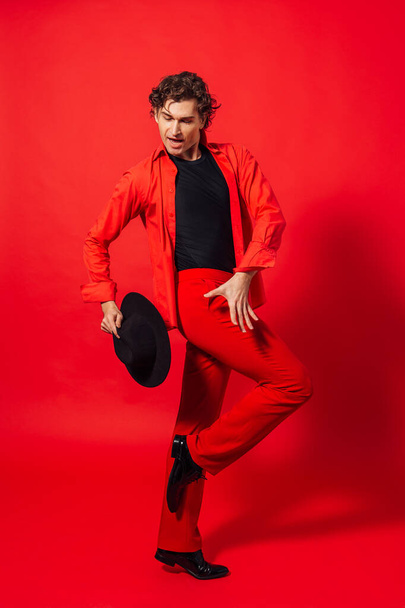 Portrait of a tall handsome man dressed in red shirt and black hat posing on the red background. - Фото, изображение