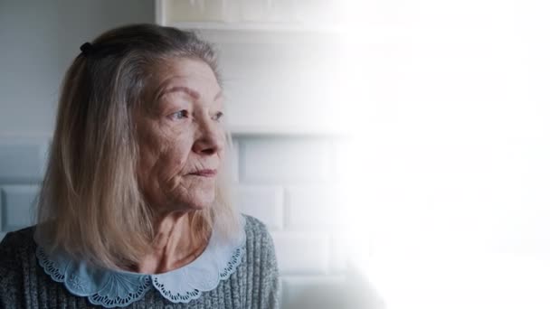 Droevige eenzame oude grijze vrouw die door het raam kijkt. Kwetsbare persoon in quarantaine - Video