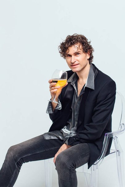 Tall handsome confident young man in a jacket sitting on the chair and holding glass of wine over white background - Фото, зображення