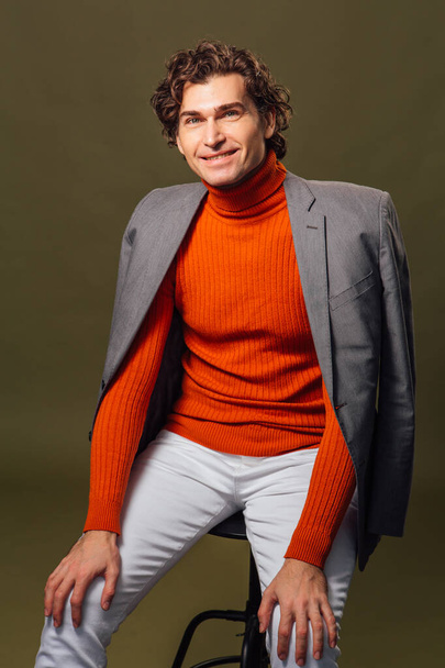 Full lengh portrait of a tall handsome man dressed in orange turtleneck, grey jacket and white jeans posing on the green background - Zdjęcie, obraz