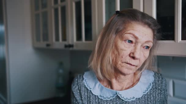 Portret van een oudere grijze dame die door het keukenraam kijkt en nadenkt. Kwetsbare eenzame mensen tijdens hevige uitbraak - Video