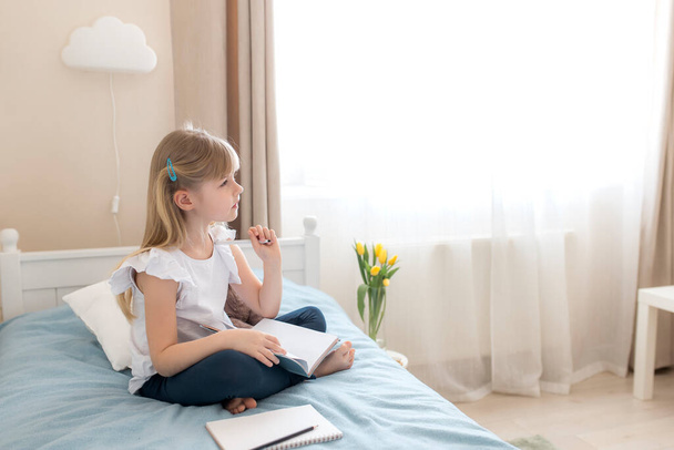Une petite fille s'assoit sur le lit dans la chambre élégante, tient un livre bleu et un stylo, faisant ses devoirs. Pense à résoudre un problème. Éducation et enseignement à domicile concept. Penser aux devoirs - Photo, image