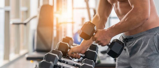 gym and fitness background banner with light flare of close up dumbbell with hand of athletic bodybuilder man lifting from dumbbell rack in gym and futness club - Fotografie, Obrázek