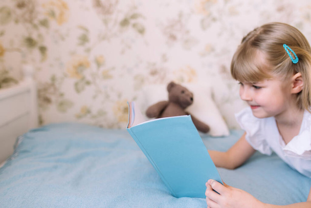 A little girl leys on the bed in the stylish bedroom and reads a blue book, doing homework. Education, home schooling concept - Photo, Image