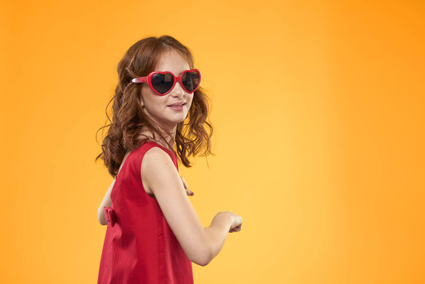 niña con gafas de sol en forma de corazón vestido rojo estudio amarillo fondo infancia - Foto, imagen