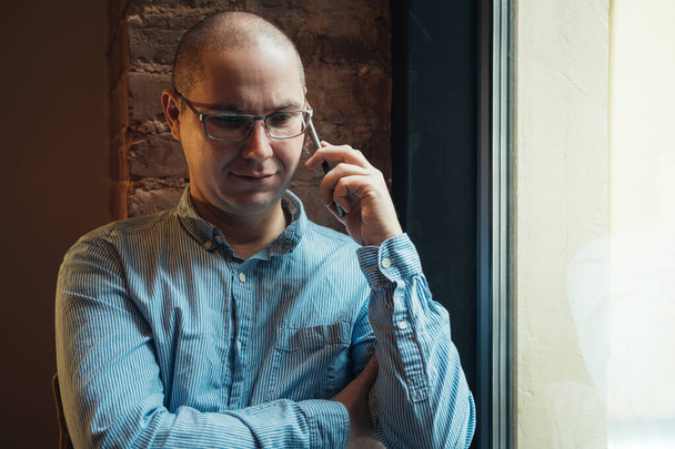 Homme en chemise bleue assis près de la fenêtre et parlant sur smartphone. Travail à domicile ou bureau de coworking moderne. - Photo, image