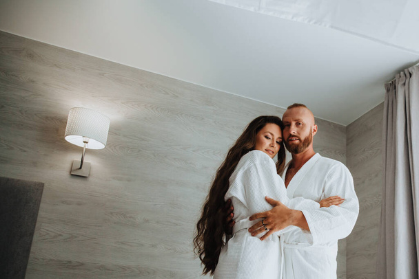 Zarte und ein schönes junges verliebtes Paar in weißen Bademänteln stehen im Luxushotel oder Schlafzimmer und umarmen sich. Gemeinsam glücklich - Foto, Bild
