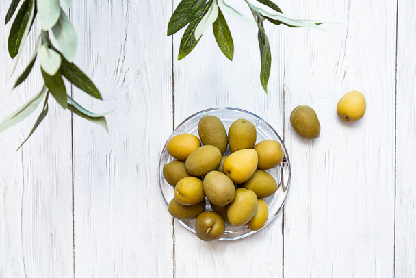 A bowl of green olives. Minimal still life of an olive branch and a glass bowl of olives on a white wooden table. Copy space - Photo, Image