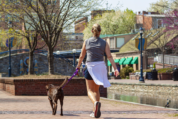 Nahaufnahme einer jungen blonden Frau, die im Park joggt, während sie ihren Hund an der Leine führt. Sie band ihre Kleider um ihre Taille. Sie trägt eine Wasserflasche bei sich. Ein malerischer Stadtpark mit Fluss. - Foto, Bild