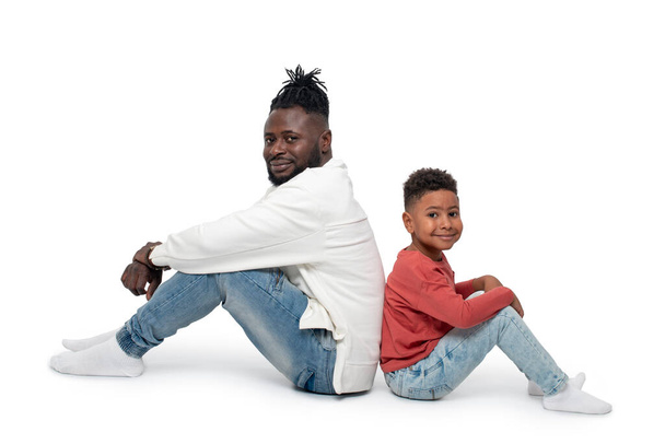 Повний портрет темношкірого африканського чоловіка і його маленького сина, що сидить на підлозі, на білому фоні у студії. - Фото, зображення