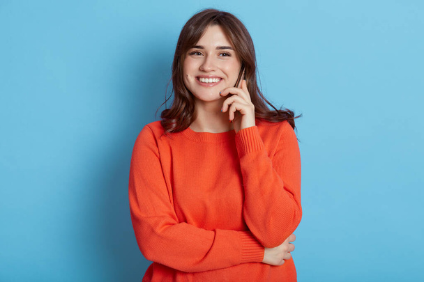 European female with happy facial expression posing with phone near ear, has pleasant conversation, hears excellent news, smiling to camera, dresses in orange jumper, isolated over blue background. - Fotografie, Obrázek