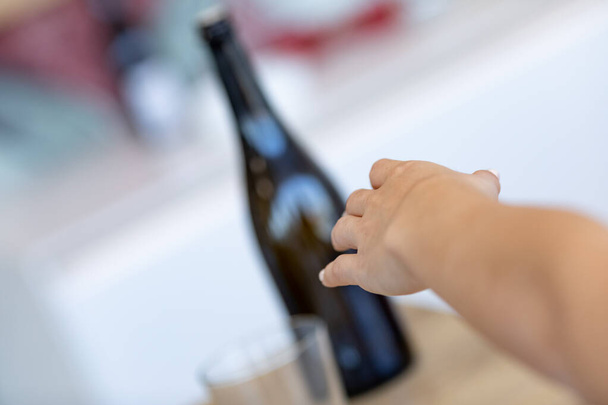 Main tendue vers une bouteille de vin sur une table à l'intérieur dans une vue d'angle incliné avec mise au point vers la main dans une perspective à la première personne - Photo, image