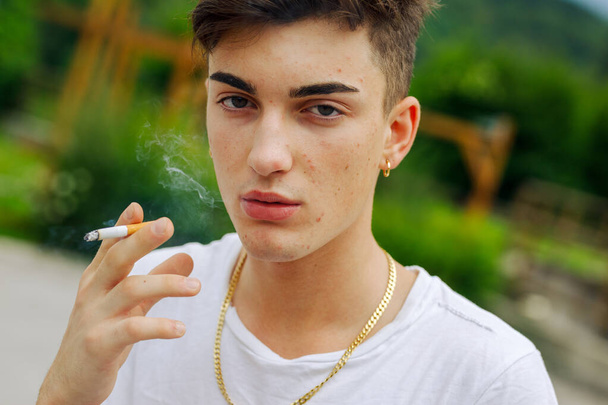Vystřižen zblízka pohled na seriózního mladého muže se zlatým řetízkem a náušnicí, jak kouří cigaretu venku a vydechuje kouř v pojetí závislosti na mládeži - Fotografie, Obrázek
