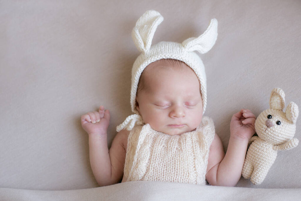Ребенок в вязаной кроличьей шляпе с ушами и кроличьей игрушкой в бежевом светлом одеяле. Весеннее фото. Пасха и дети. Счастливого материнства. Счастливой Пасхи  - Фото, изображение