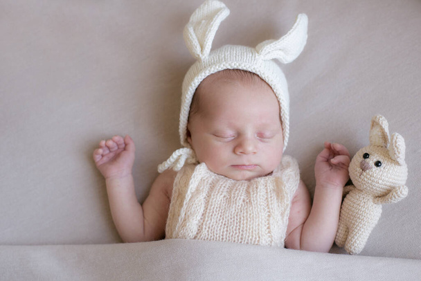 Kulakları olan örülü tavşan şapkalı bir bebek ve bej renkli battaniyeli tavşan oyuncağı. Bahar fotoğrafı. Paskalya ve çocuklar. Mutlu annelikler. Mutlu Paskalyalar  - Fotoğraf, Görsel