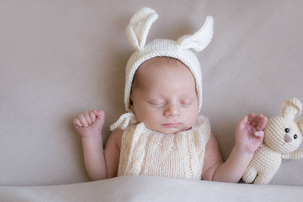 Baby in a knitted rabbit hat with ears and a bunny toy in a beige light blanket. Spring photo. Easter and children. Happy motherhood. Happy easter  - Фото, изображение
