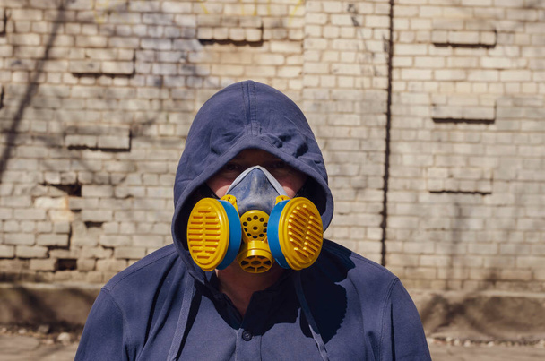 Portrait of an adult unrecognizable man wearing a respirator and hood on against a brick wall. Middle-aged man wearing an industrial blue-and-yellow respirator looks into the camera. Outside. Daytime - Zdjęcie, obraz