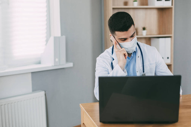 Jeune médecin masculin masqué travaillant sur un ordinateur portable dans son bureau - Photo, image
