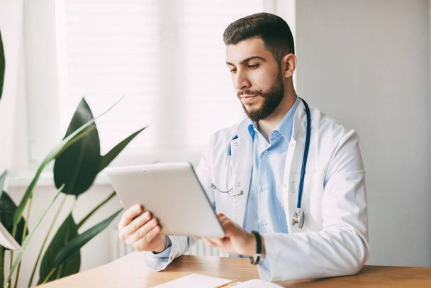 Mužský lékař orientálního vzhledu soustředěně pracuje na tabletu, sedí u stolu ve své kanceláři. - Fotografie, Obrázek