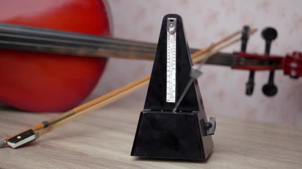 Egy fekete vintage metronóm üti a ritmust, miközben az asztalon áll, mögötte egy cselló, egy közeli felvétel Hangszerkoncepció - Felvétel, videó