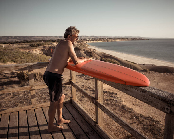 Attractive Australian mature man surfer with cool vintage surfboard on beach at sunset. Senior adult happy to be Back to surf . Outdoors sports adventure, active older people and retirement lifestyle. - Фото, изображение