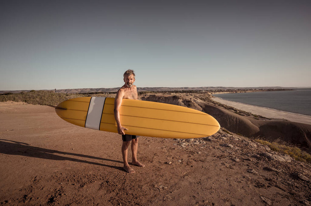 Attractive Australian mature man surfer with cool vintage surfboard on beach at sunset. Senior adult happy to be Back to surf . Outdoors sports adventure, active older people and retirement lifestyle. - Φωτογραφία, εικόνα