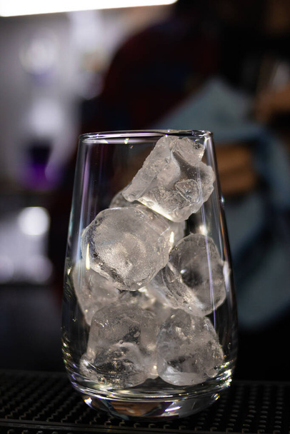 A vertical shot of a glass full of ice cubs on blurred bar background - Foto, Imagem