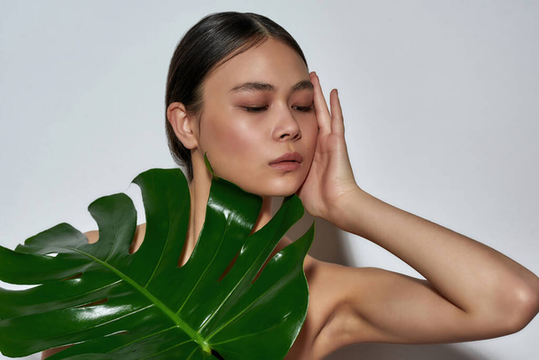 Portrait of a young magnificent half-naked asian girl in a black top with a monstera leaf and gently touching face - Foto, afbeelding