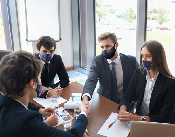 Businessman in preventive mask shaking hands to seal a deal with his partner and colleagues who also wear preventive masks in office - Фото, зображення